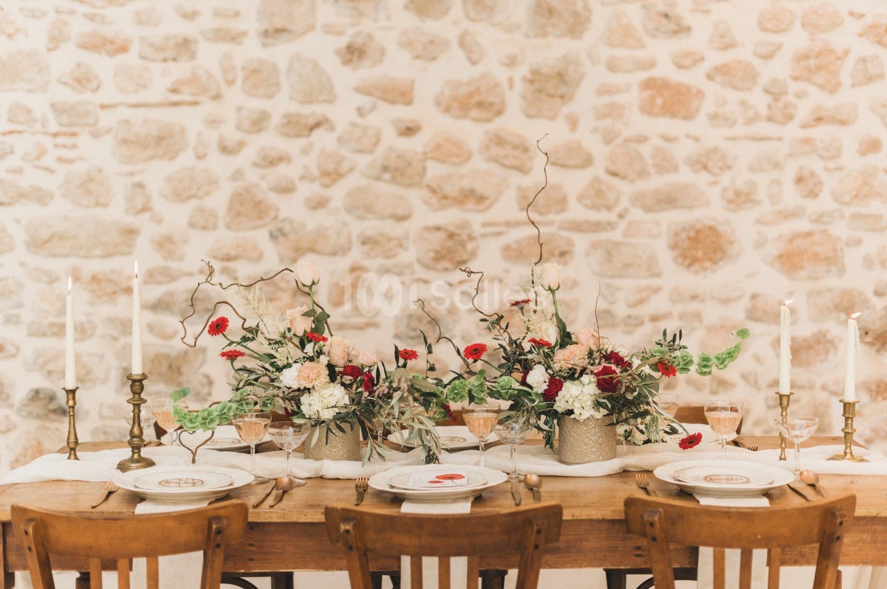 Table en bois décorée de fleurs colorées, bougies et vaisselle élégante, devant un mur en pierre apparente.