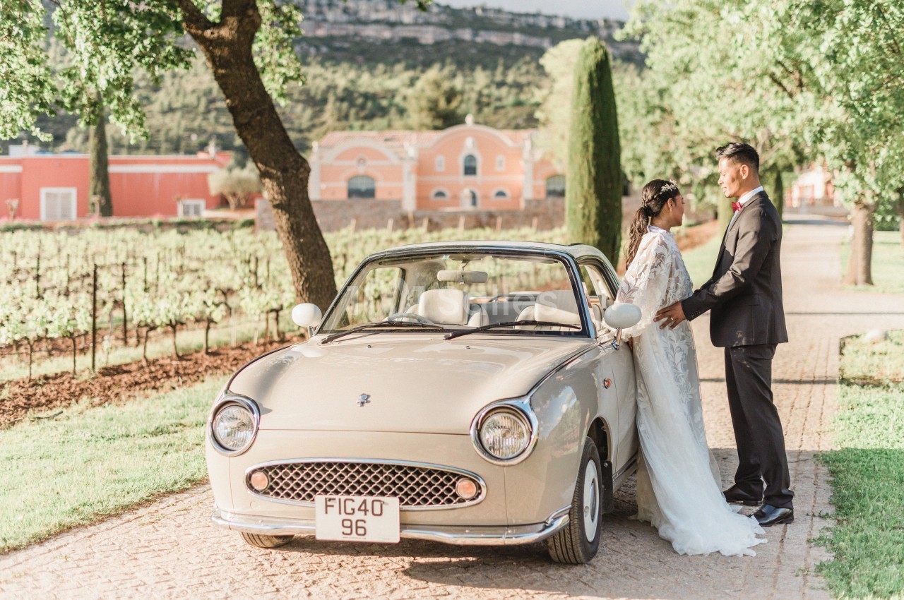 Un couple en tenue de mariage se tient près d'une voiture vintage beige, dans une allée bordée d'arbres et de vignes.