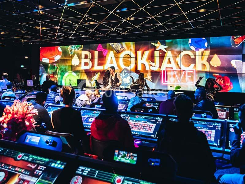 Salle de casino animée avec des joueurs devant des écrans individuels et un écran géant affichant ’Blackjack Live’.