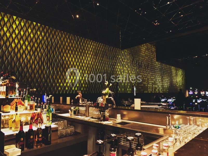 Bar moderne avec comptoir en métal, bouteilles alignées et mur illuminé par un motif géométrique doré.
