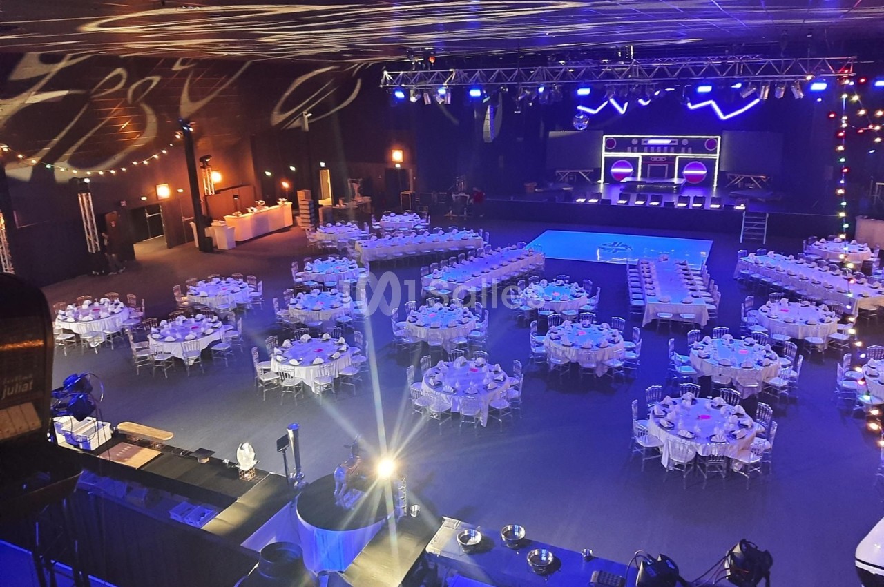 Salle de réception avec tables rondes dressées, éclairages colorés et scène équipée pour un événement.