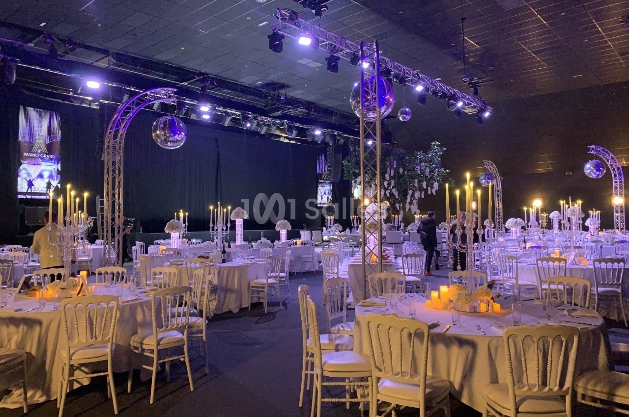 Salle de réception décorée avec des tables rondes, nappes blanches, chandeliers et éclairage tamisé.