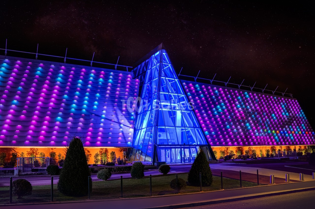 Façade illuminée d'un bâtiment moderne avec des motifs colorés et une entrée vitrée triangulaire, de nuit.