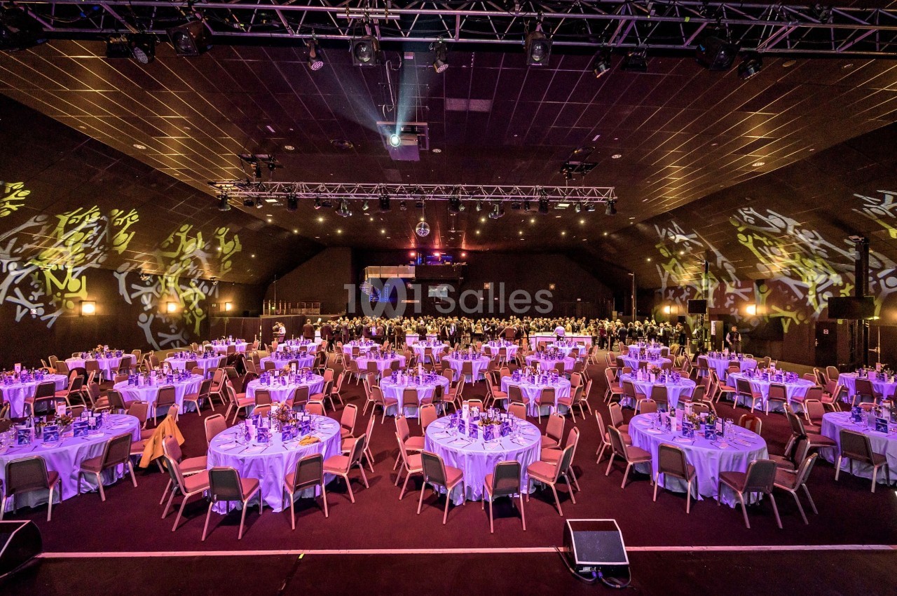 Salle de réception avec de nombreuses tables rondes dressées pour un événement, éclairée par des lumières colorées.