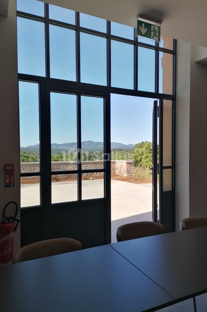 Vue sur un paysage extérieur depuis une salle avec une grande baie vitrée ouverte, table et chaises au premier plan.