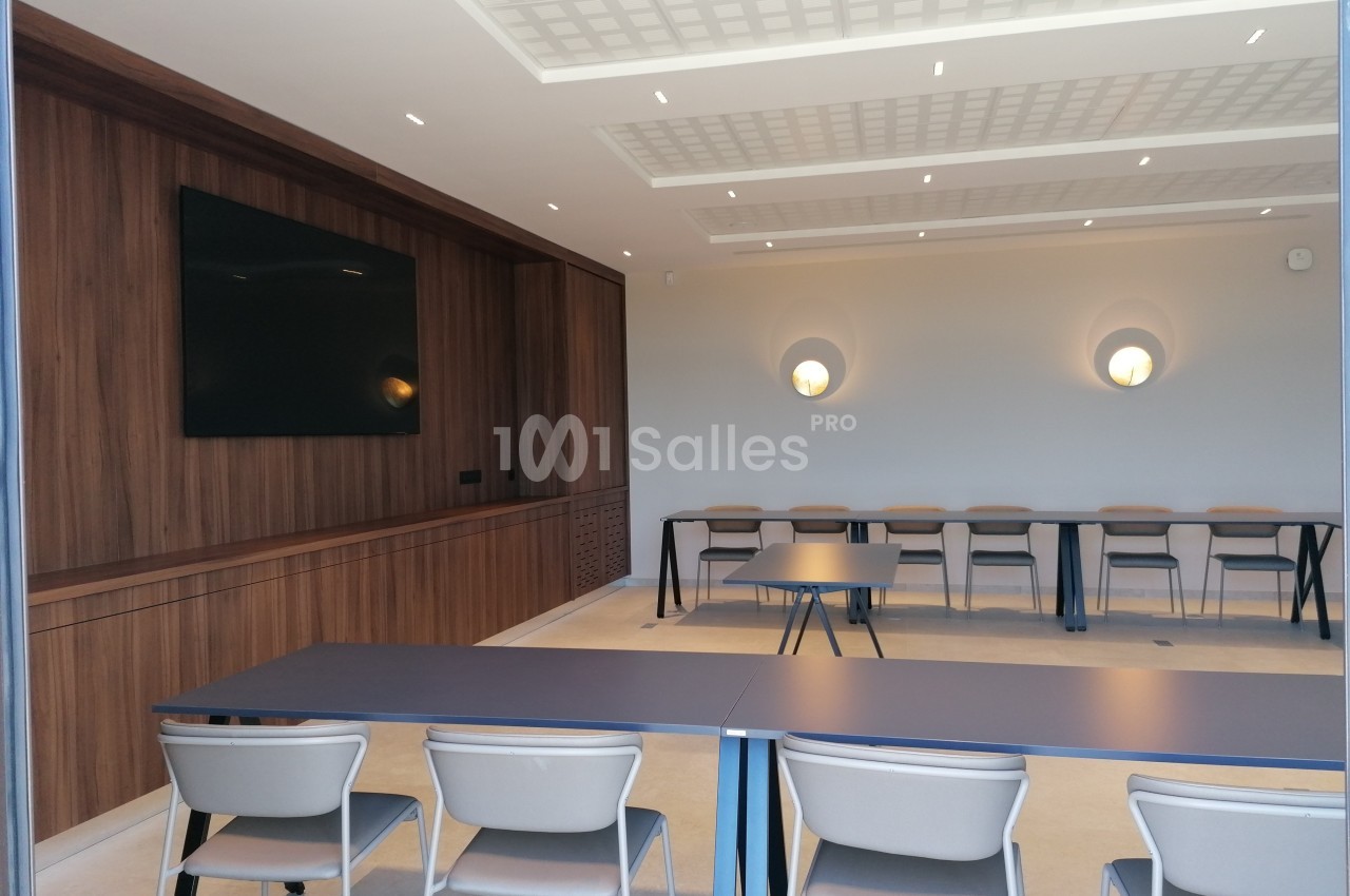 Salle de réunion moderne avec tables, chaises alignées et écran mural, éclairée par des appliques murales.