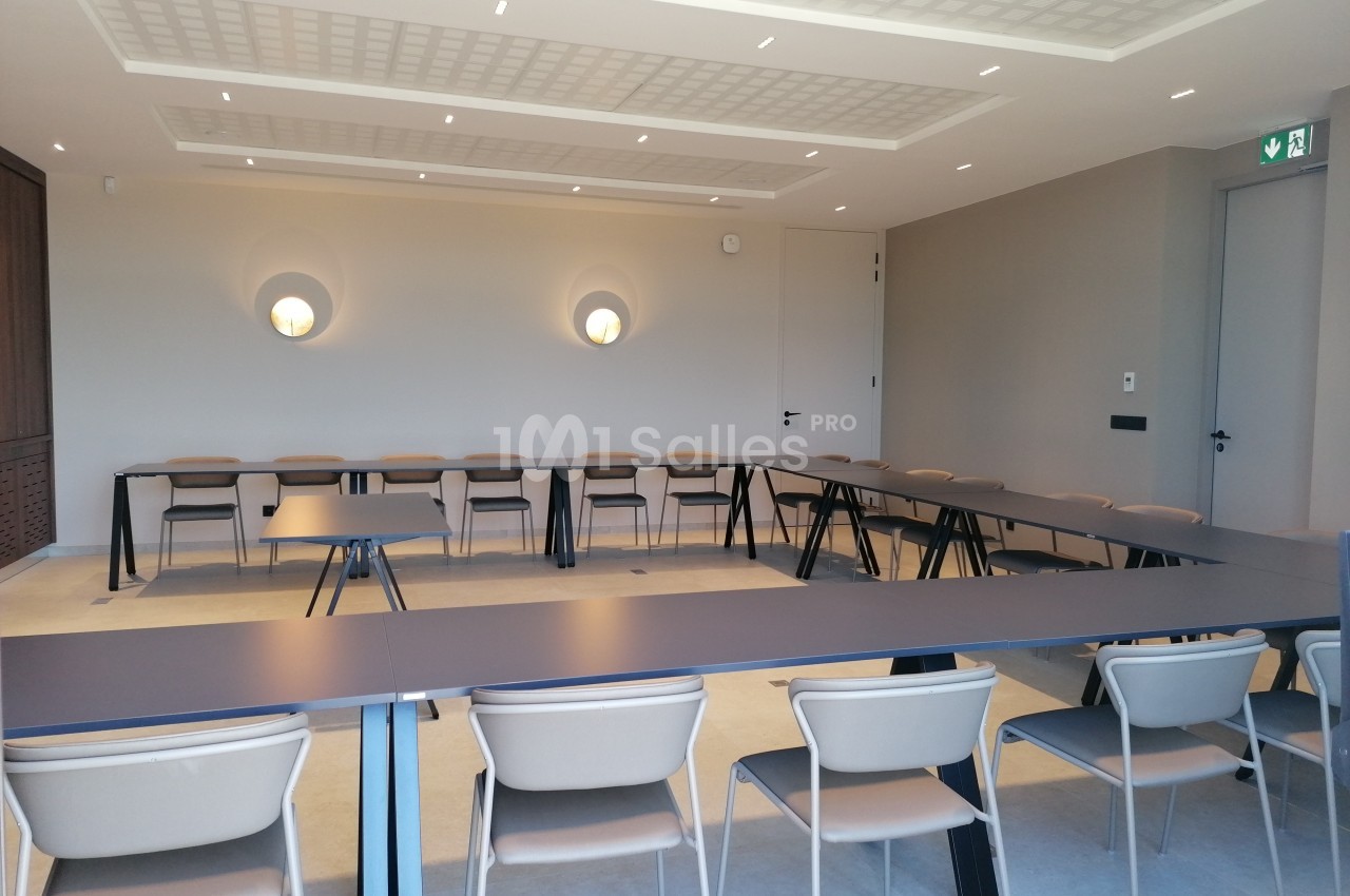 Salle de réunion lumineuse avec tables disposées en U et chaises beige clair, murs neutres et éclairage mural.
