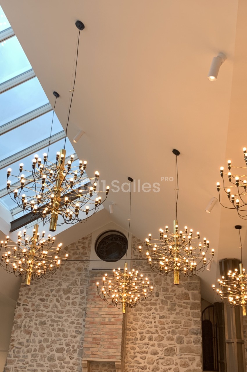 Plafond avec lustres dorés suspendus, mur en pierre apparente et verrière laissant entrer la lumière naturelle.