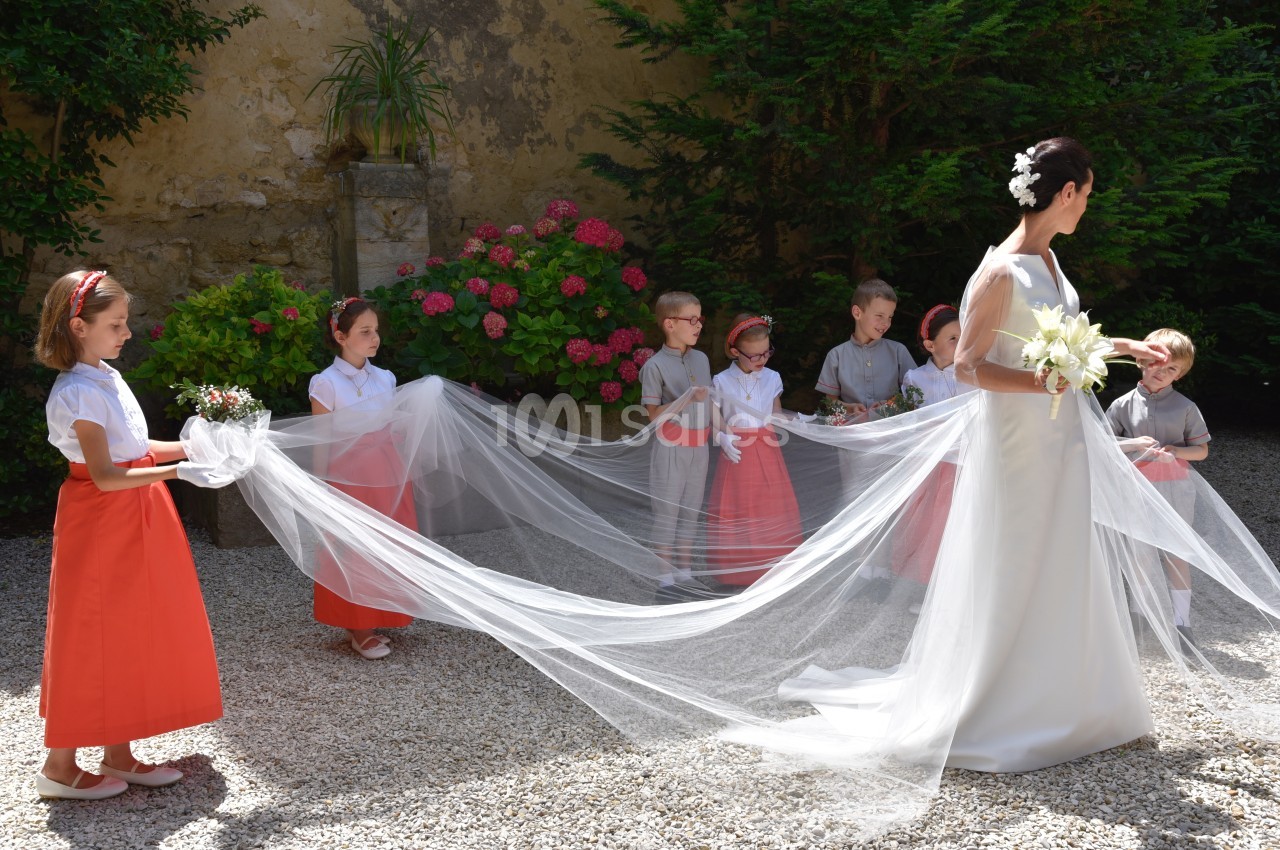 Une mariée en robe blanche avec un long voile tenu par des enfants d'honneur dans un jardin fleuri.