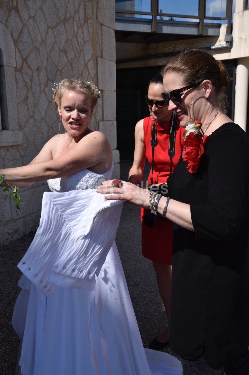 Une mariée ajuste sa robe blanche avec l'aide de deux femmes à l'extérieur d'un bâtiment en pierre.