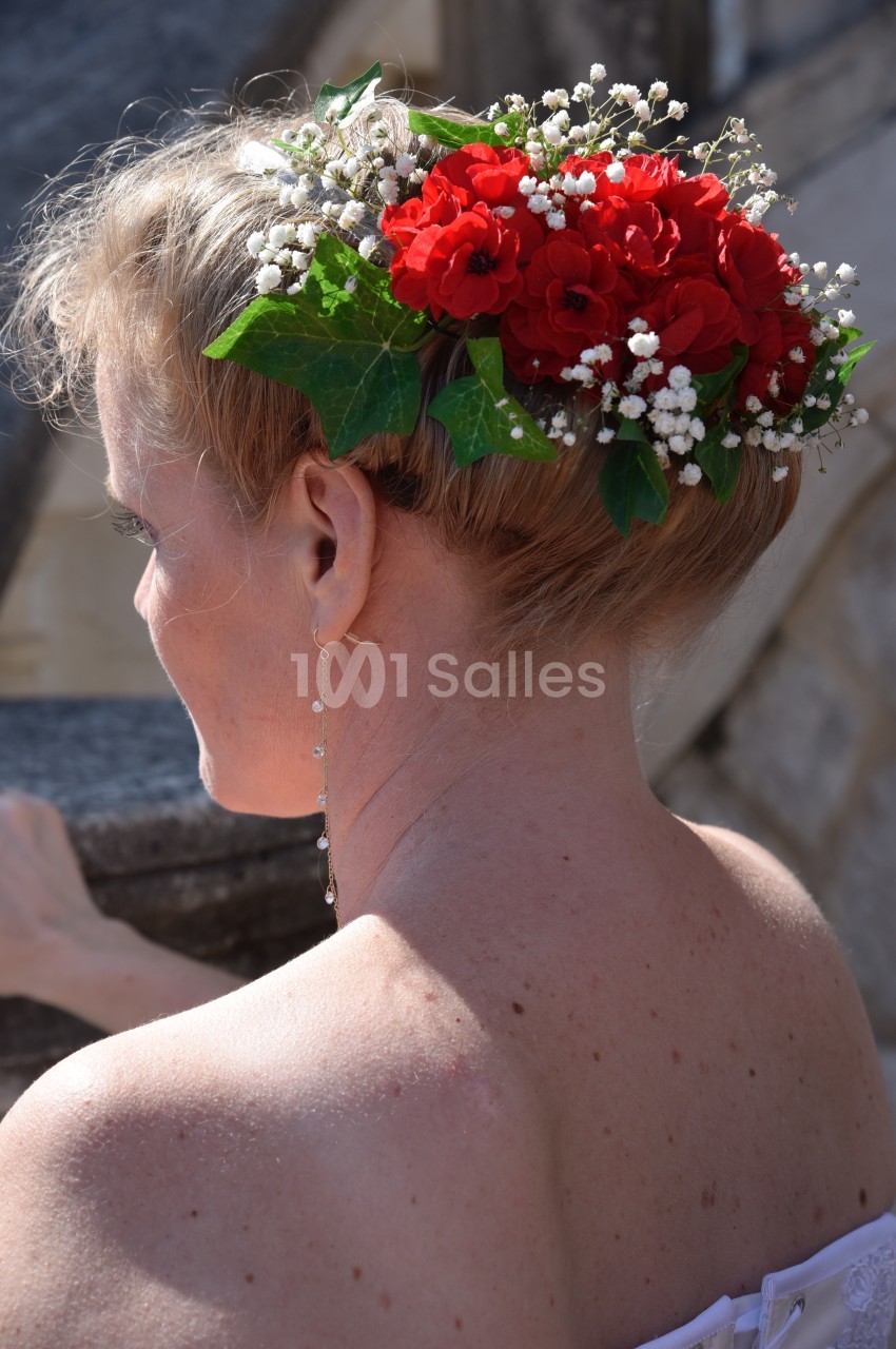 Coiffure de mariage avec chignon orné de fleurs rouges, gypsophile et feuillage, vue de dos en extérieur.