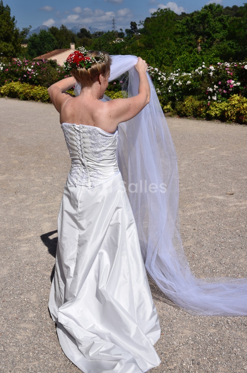 Une femme en robe de mariée blanche ajuste un long voile dans un jardin fleuri sous un ciel ensoleillé.