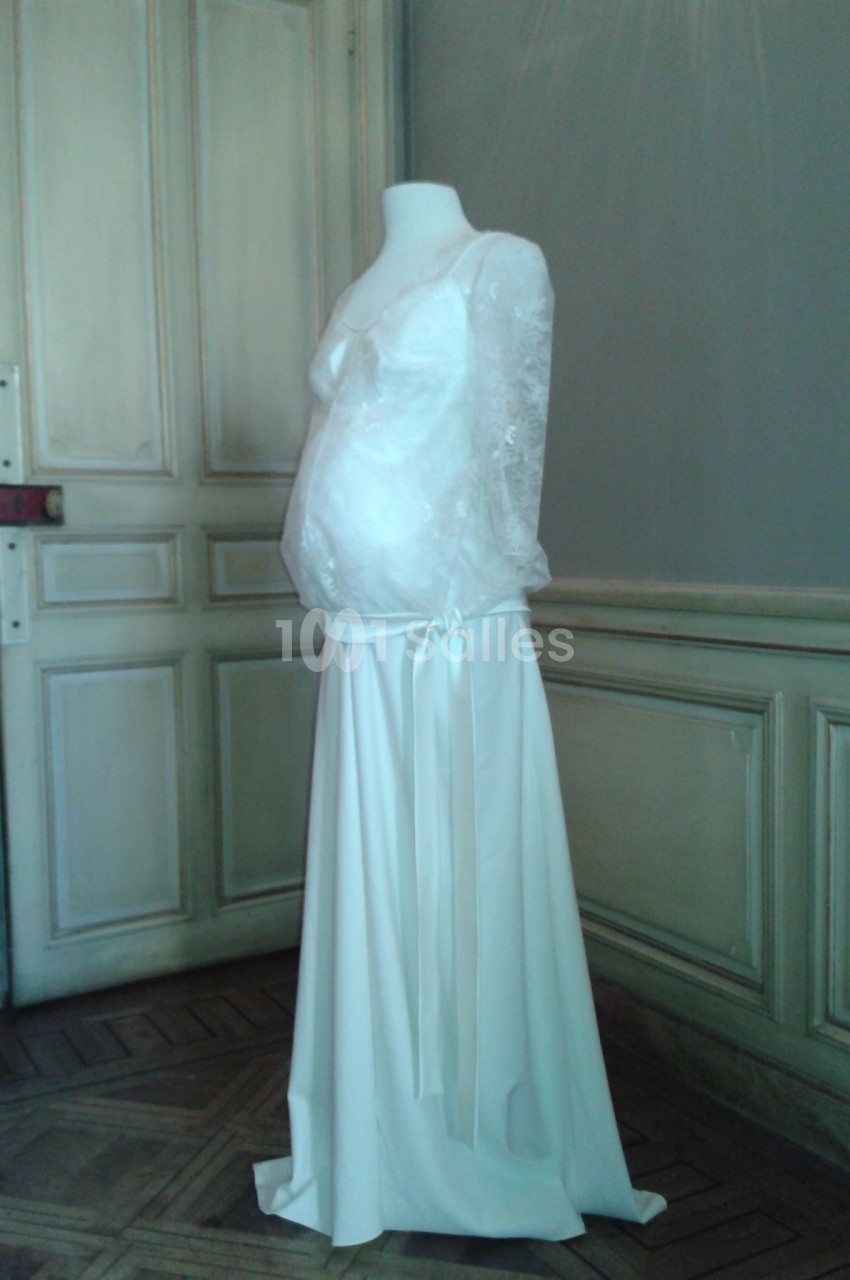 Robe de mariée blanche avec manches longues en dentelle, présentée sur un mannequin dans une pièce au décor classique.