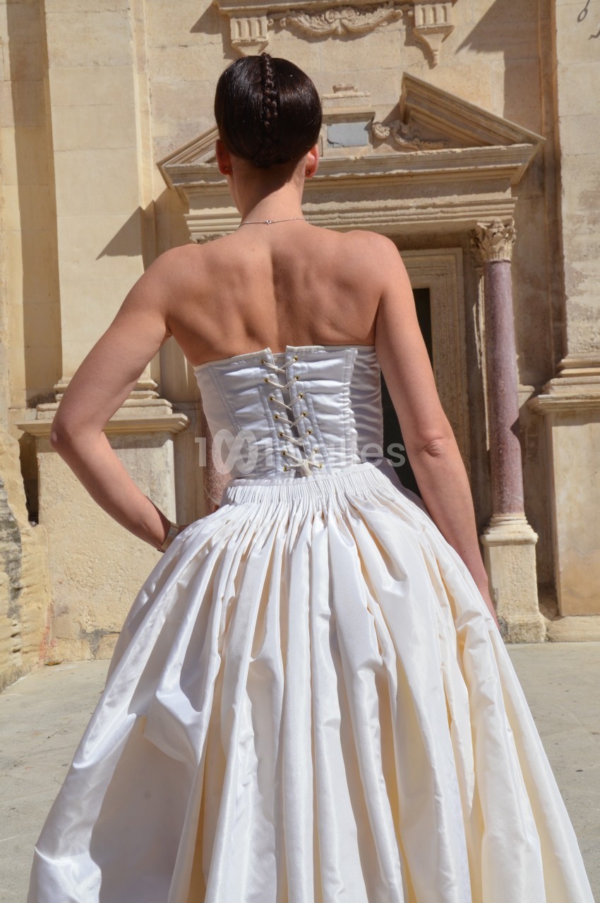 Femme de dos portant une robe blanche corsetée devant un bâtiment ancien en pierre.