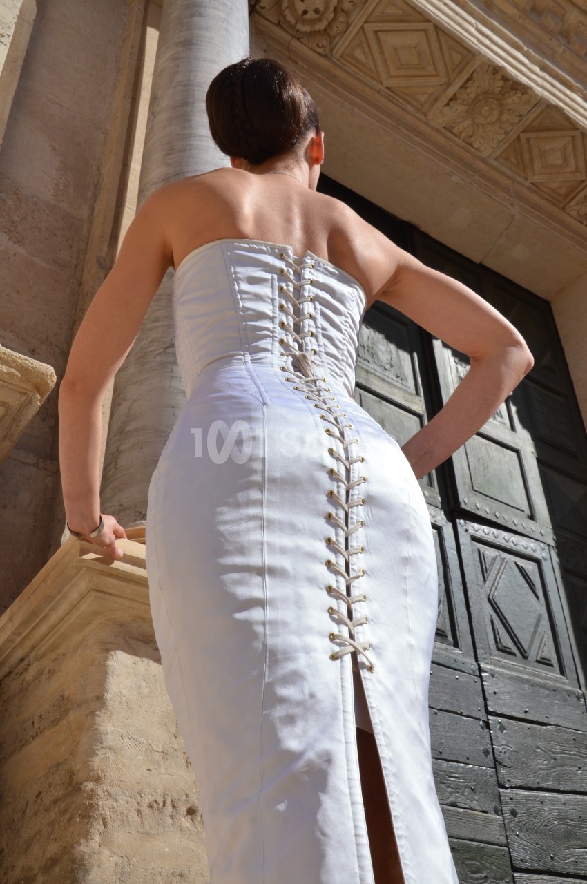 Femme en robe blanche ajustée, vue de dos, devant une porte en bois et une colonne en pierre.