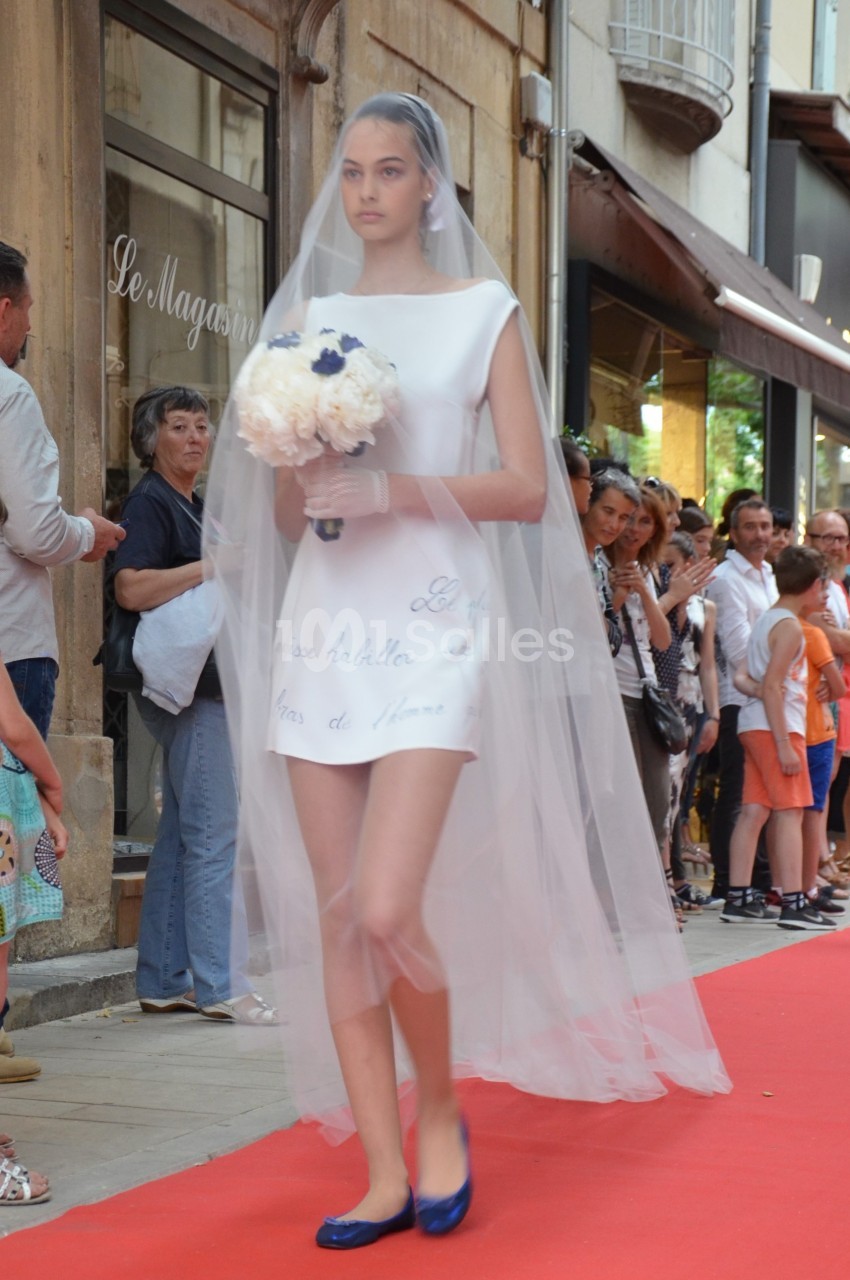 Une femme en robe blanche courte et voile de mariée marche sur un tapis rouge, tenant un bouquet de fleurs.