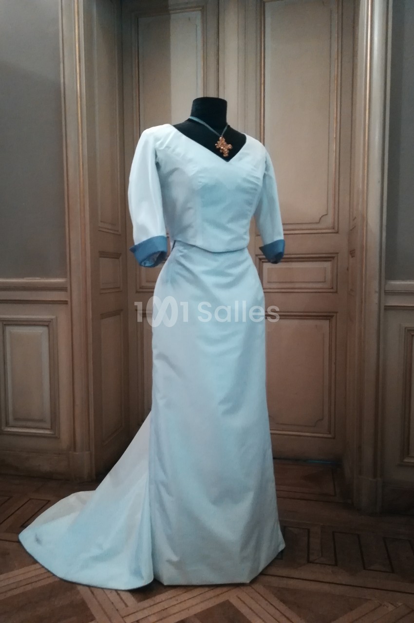 Robe longue blanche avec manches trois-quarts et détails bleus, présentée sur un mannequin dans une pièce élégante.