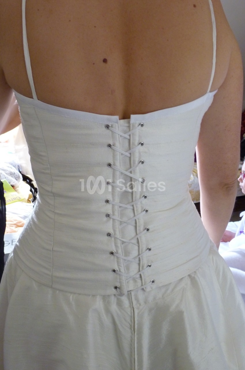 Dos d'une femme portant une robe de mariée blanche avec un corset lacé, vue dans un environnement intérieur.
