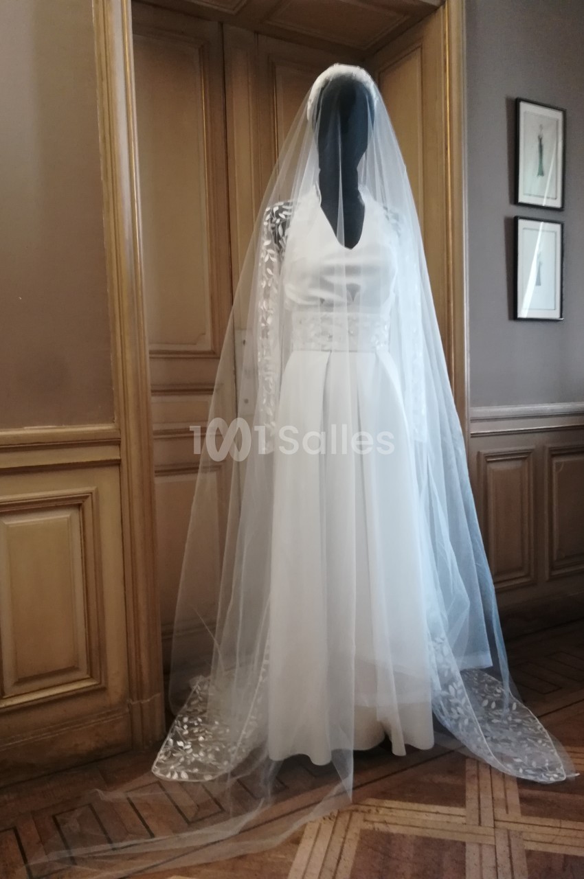 Mannequin portant une robe de mariée blanche avec un long voile, placé devant une porte en bois dans une pièce élégante.