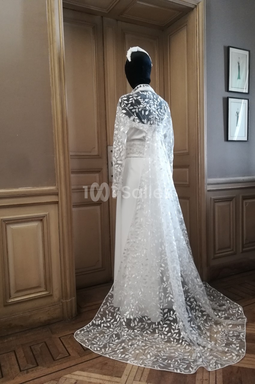 Mannequin vêtu d'une robe de mariée blanche avec une longue cape en dentelle florale, dans une pièce élégante.