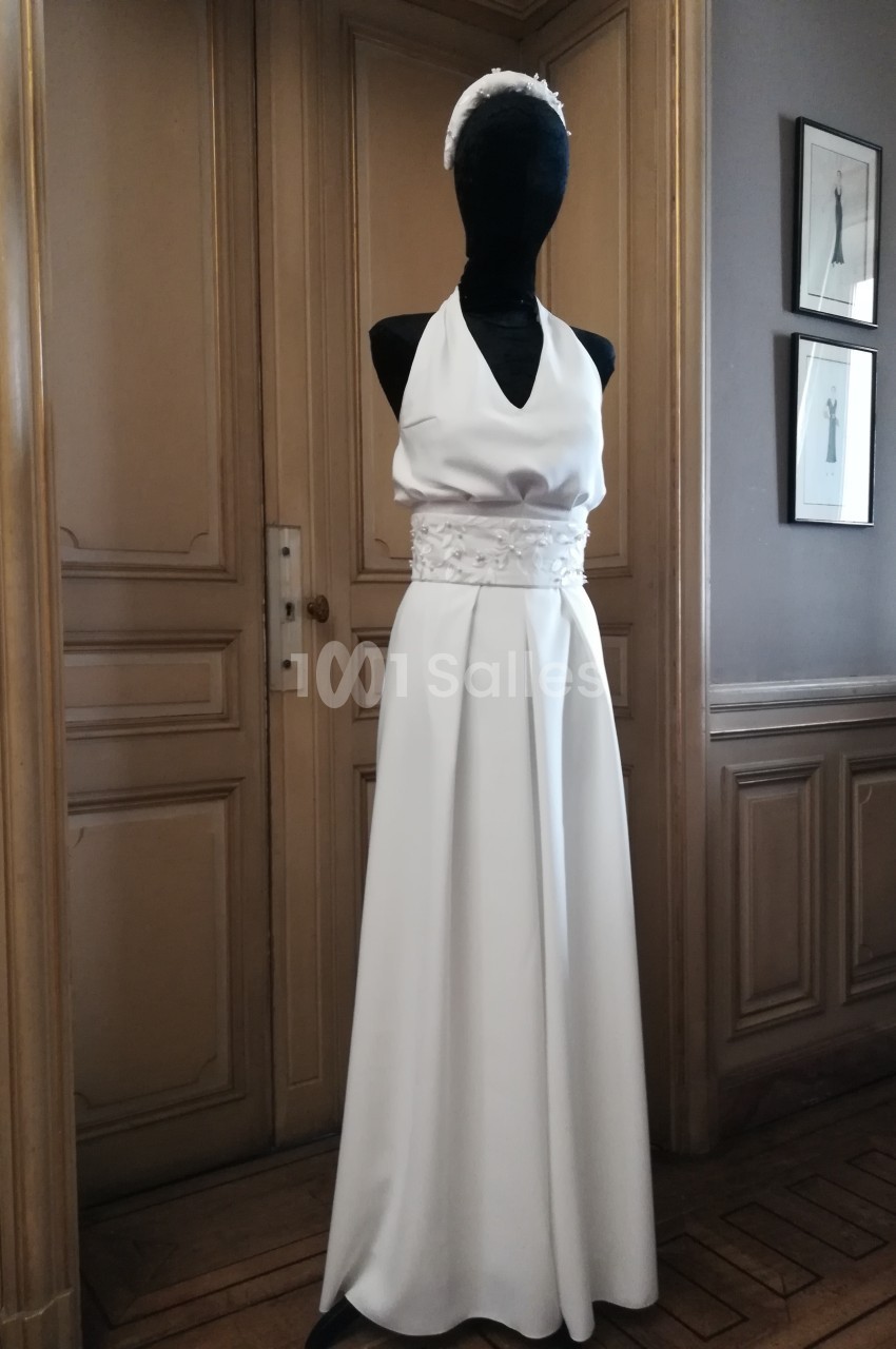 Mannequin portant une robe de mariée blanche sans manches avec ceinture ornée, dans une pièce au décor classique.