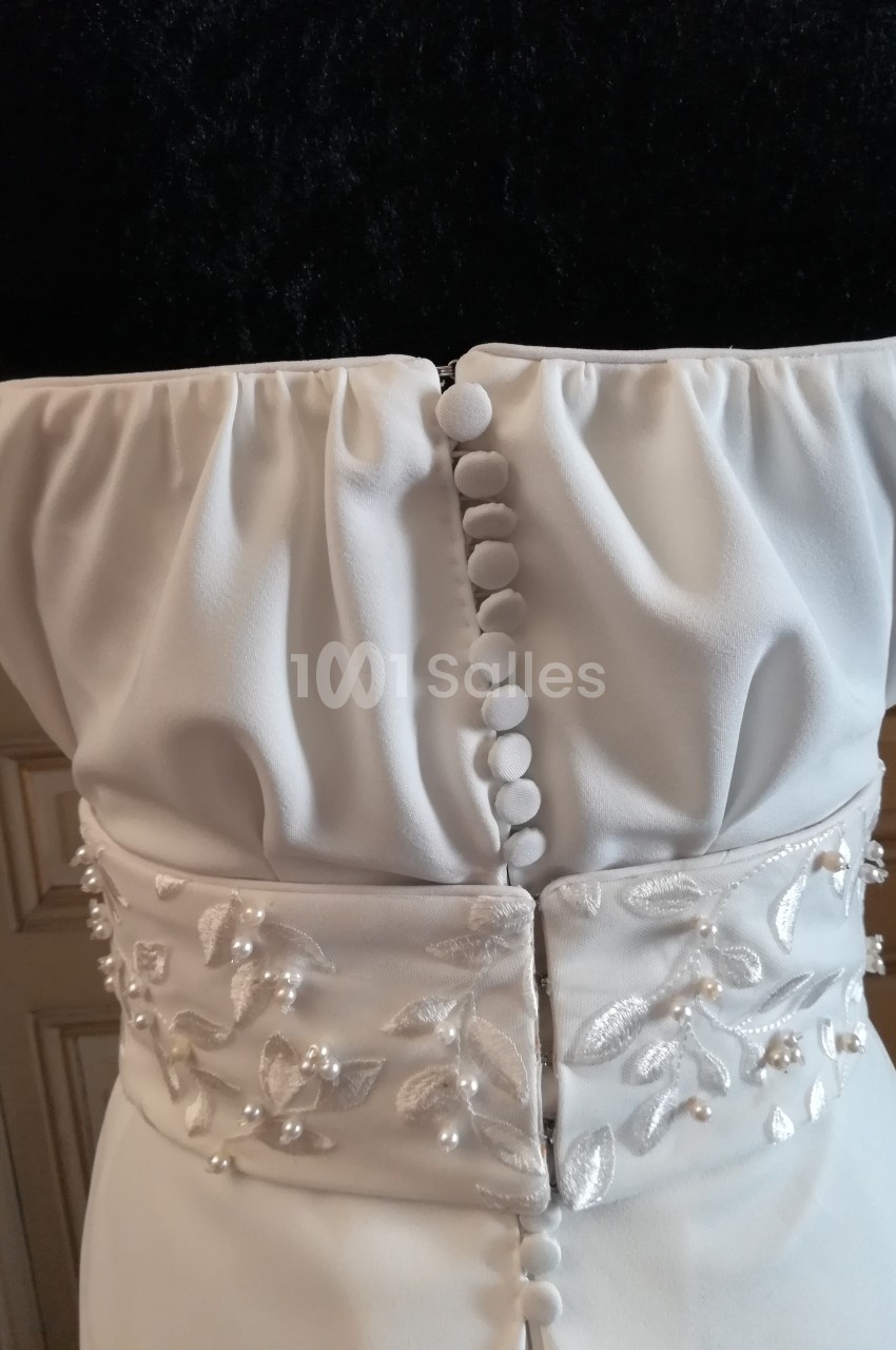 Dos d'une robe blanche avec boutons recouvrant la fermeture et ceinture ornée de broderies florales et perles.