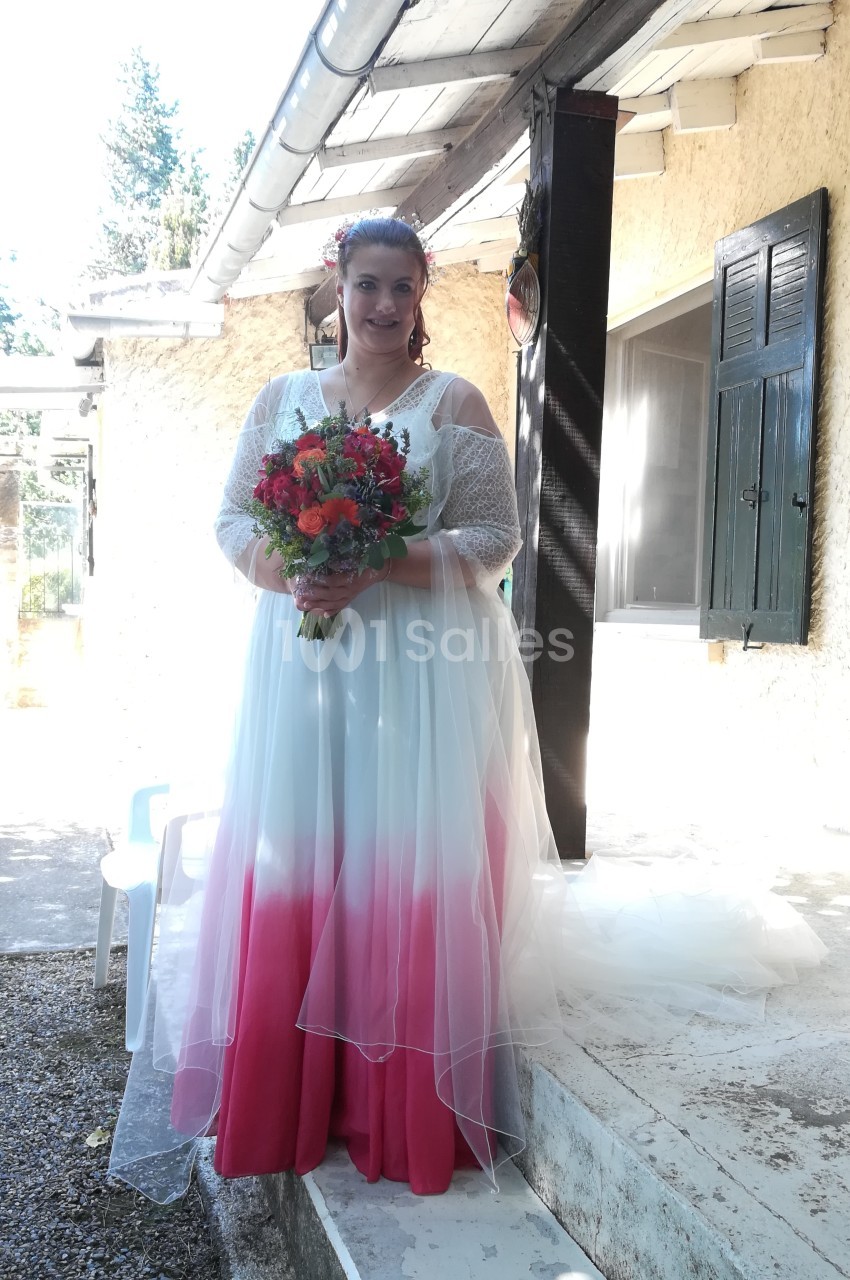 Femme en robe de mariée blanche et rose tenant un bouquet coloré, debout devant une maison ensoleillée.