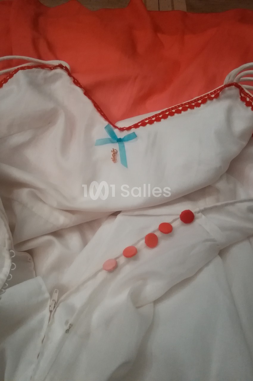Robe blanche avec bordures rouges, ornée d'un nœud bleu et de boutons rouges, posée sur un tissu orange.
