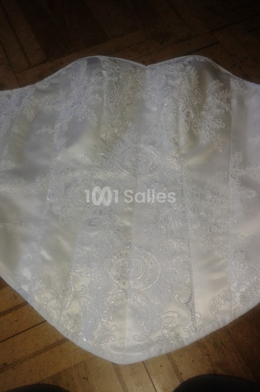 Corset blanc en satin orné de dentelle délicate, posé sur un sol en parquet clair.