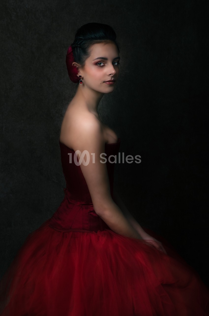 Femme en robe rouge élégante, assise de profil dans un éclairage tamisé sur fond sombre.