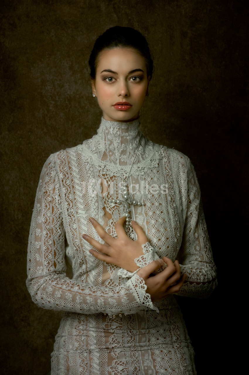 Femme en robe en dentelle blanche, posant devant un fond sombre avec les mains croisées sur le buste.