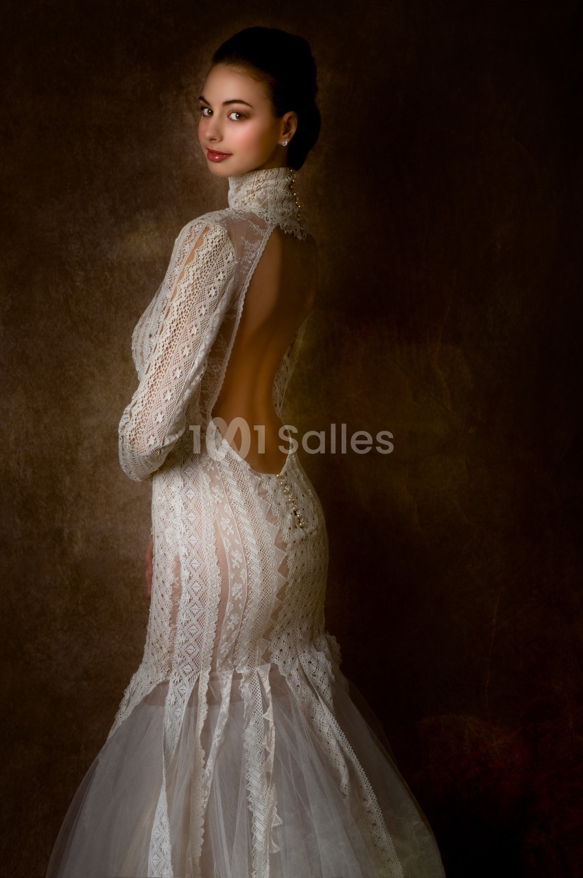 Femme portant une robe de mariée en dentelle avec un dos ouvert, posant devant un fond sombre.