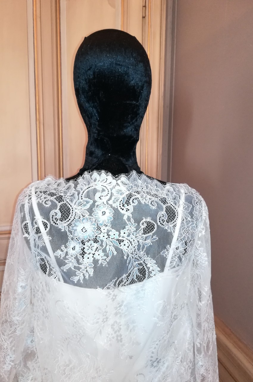 Mannequin vêtu d'une robe en dentelle blanche avec un voile noir couvrant la tête, devant une porte en bois clair.