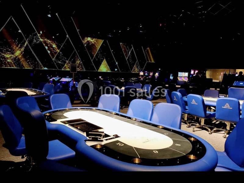 Salle de casino avec tables de jeu et chaises bleues, éclairage tamisé et écrans lumineux en arrière-plan.