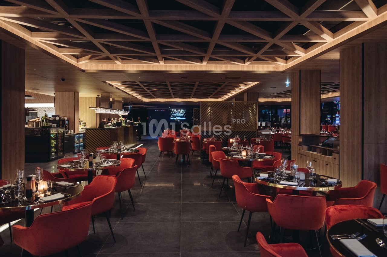 Salle de restaurant élégante avec des tables dressées, des chaises rouges et un éclairage tamisé sous un plafond à motifs.
