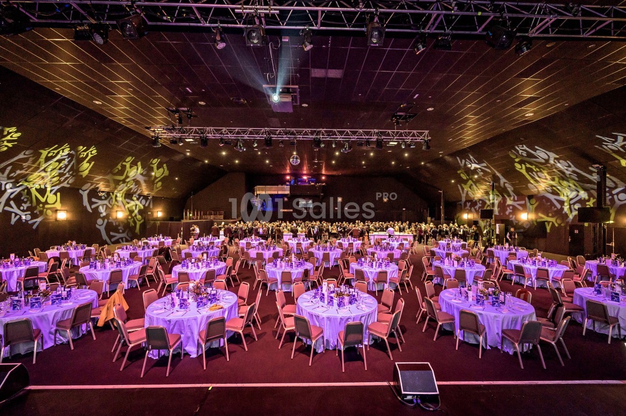 Salle de réception avec de nombreuses tables rondes dressées pour un événement, éclairée par des lumières colorées.