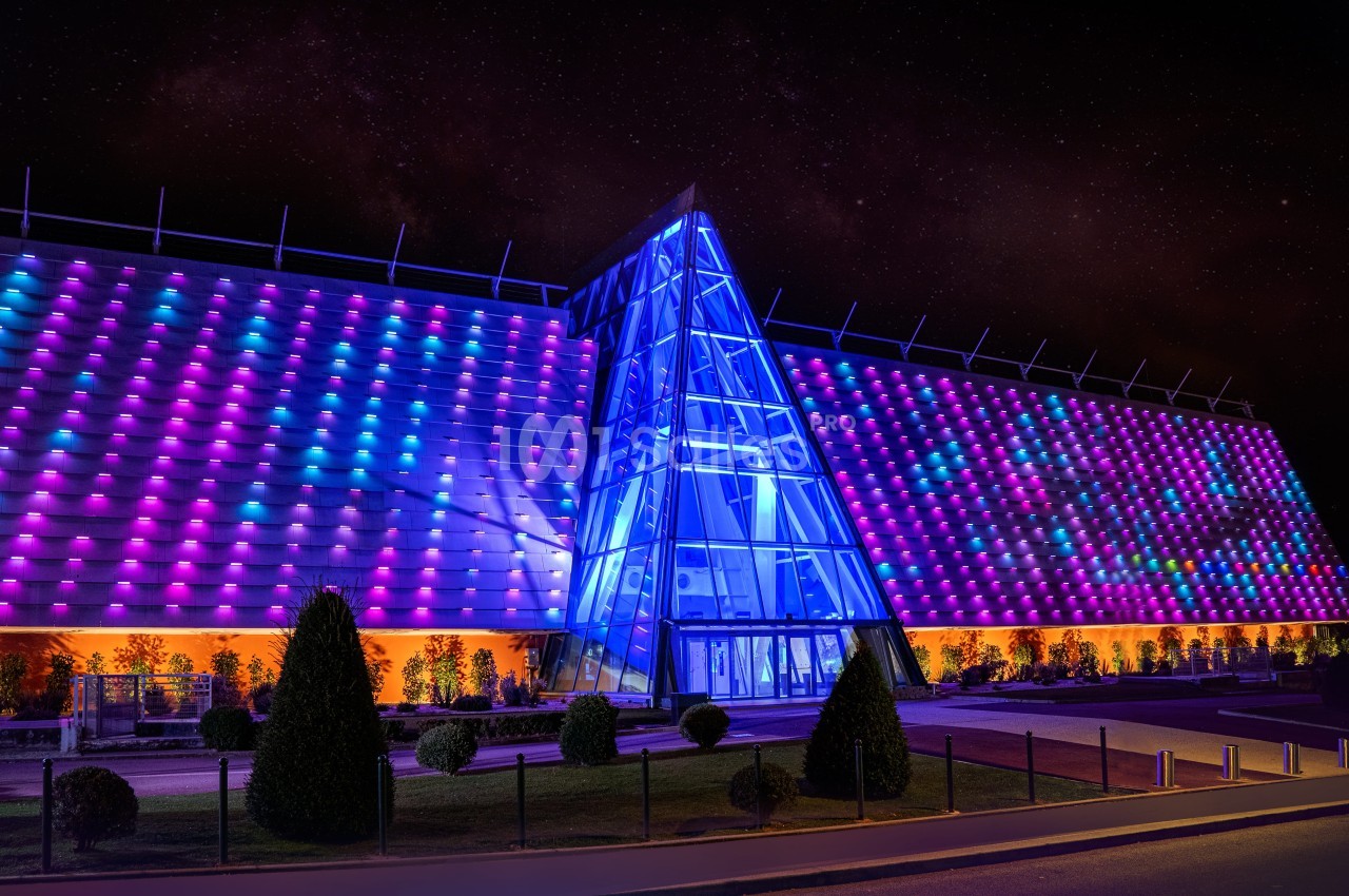 Façade illuminée d'un bâtiment moderne avec des motifs colorés et une entrée vitrée triangulaire, de nuit.