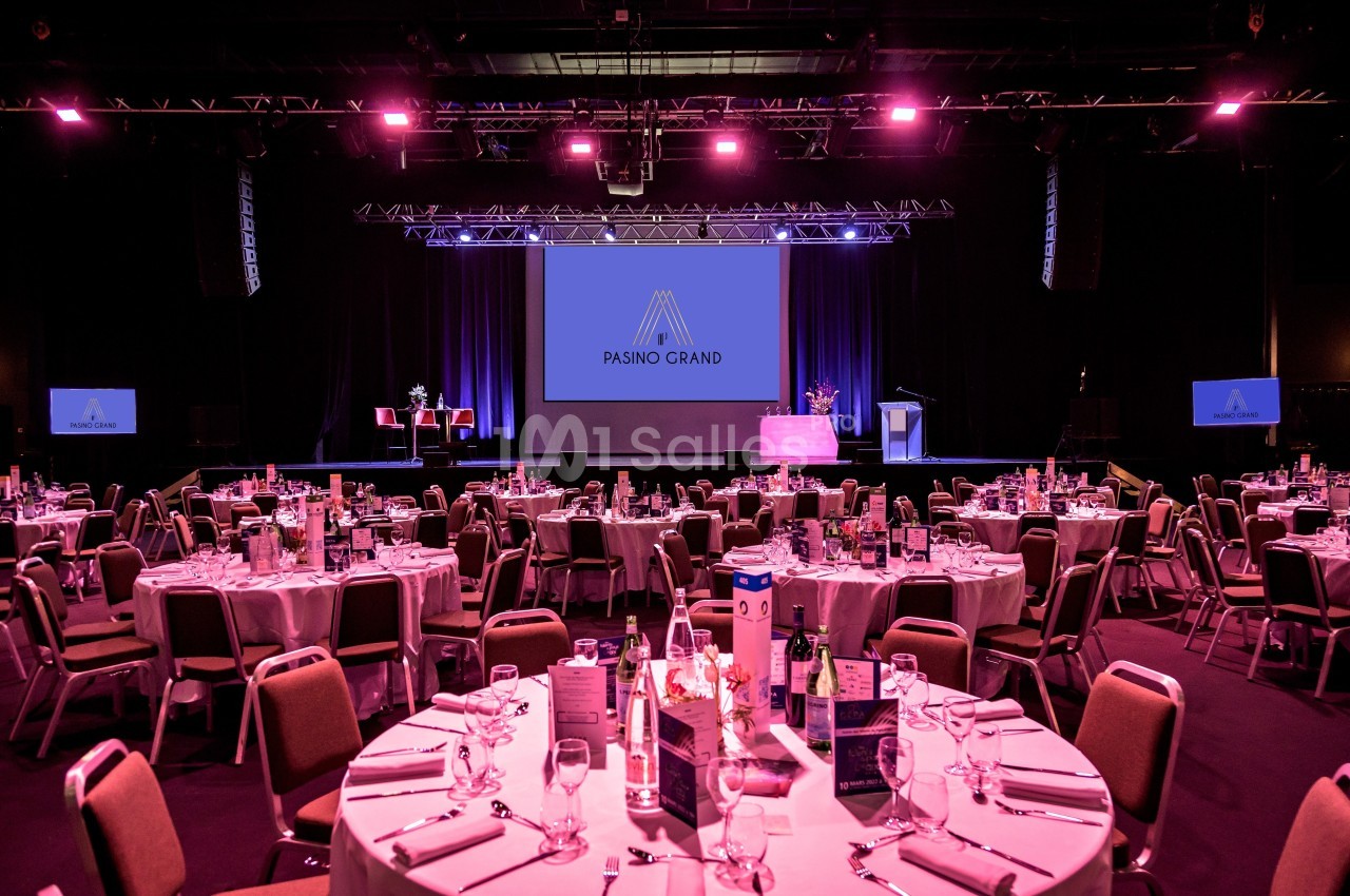 Salle de réception avec tables dressées et scène éclairée, équipée d'écrans affichant le logo ’Pasino Grand’.