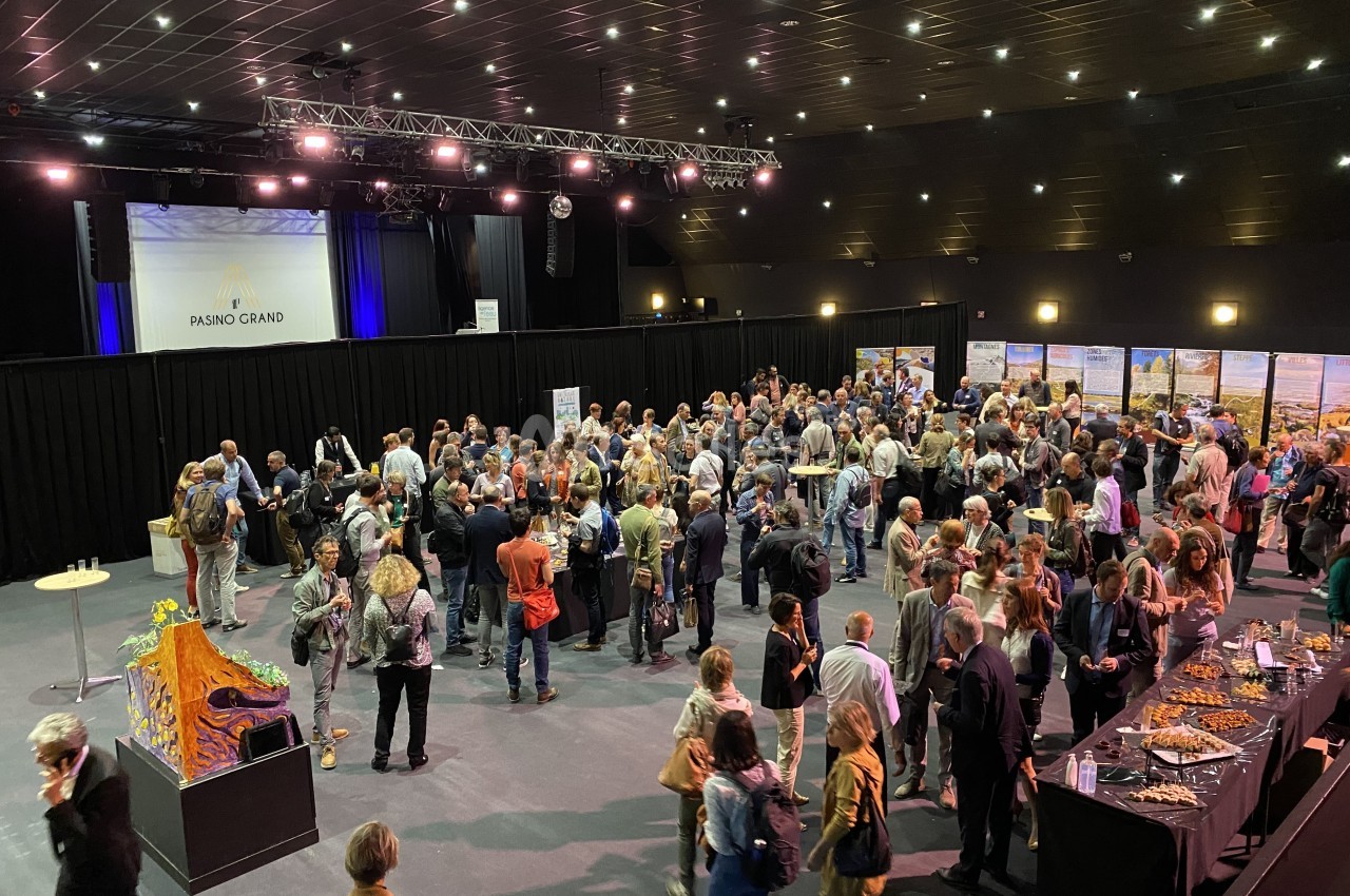 Groupe de personnes rassemblées dans une salle de conférence avec stands d'exposition et buffet.