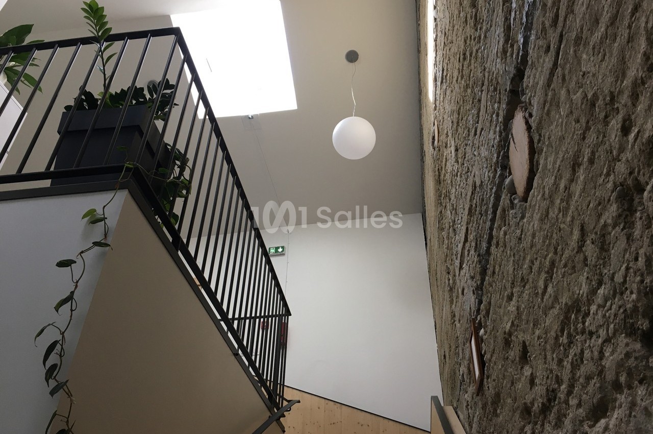 Escalier intérieur avec garde-corps en métal, mur en pierre apparente et éclairage naturel provenant d'un puits de lumière.