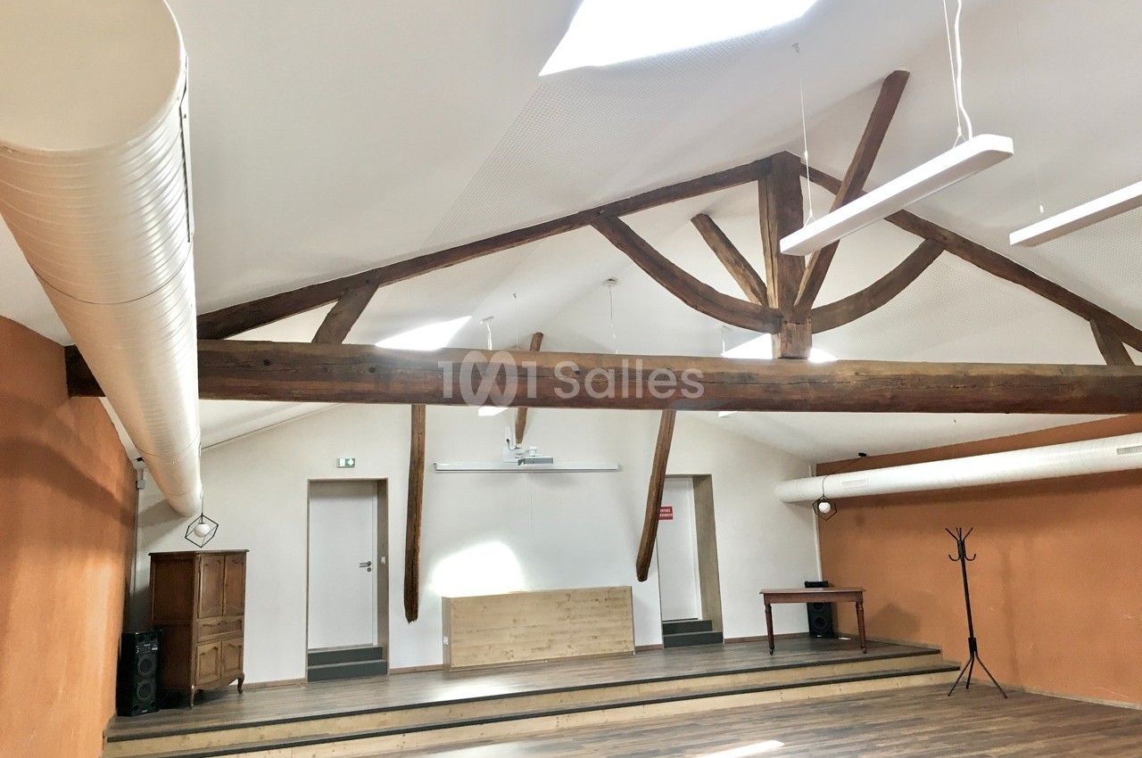 Salle lumineuse avec poutres apparentes, parquet au sol, murs orange et blancs, équipée d’un projecteur et de mobilier…