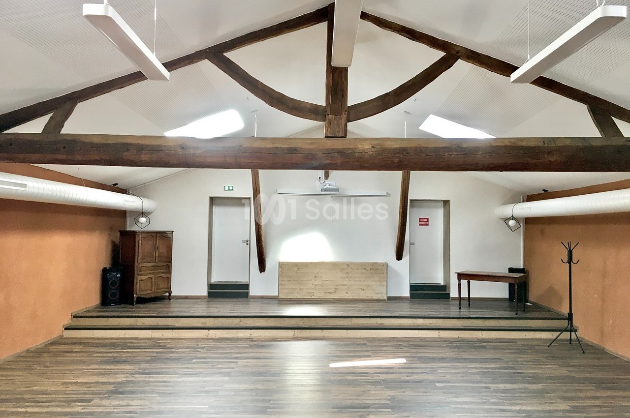 Salle lumineuse avec poutres apparentes, parquet en bois, estrade, mobilier minimaliste et éclairage suspendu.