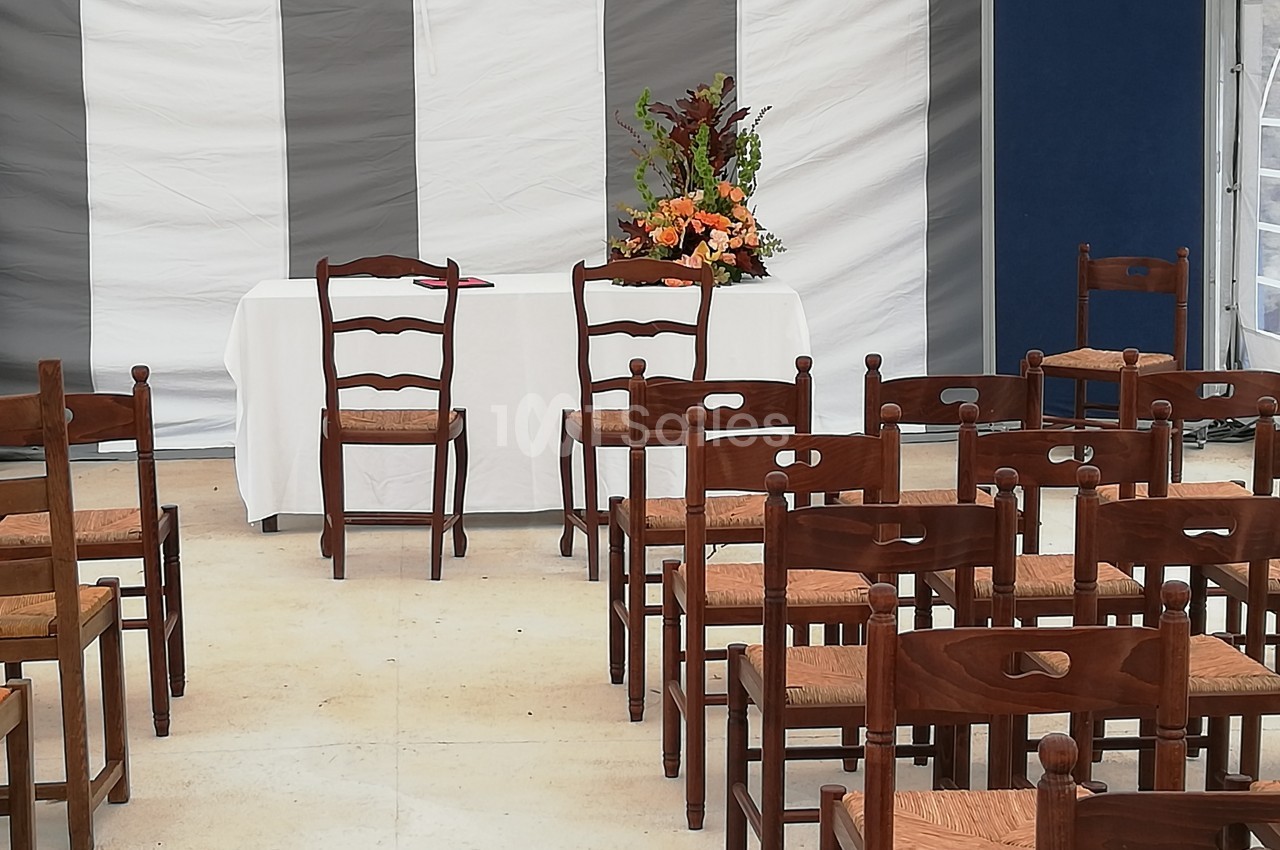 Salle aménagée avec des chaises en bois face à une table blanche décorée de fleurs, dans un espace couvert.