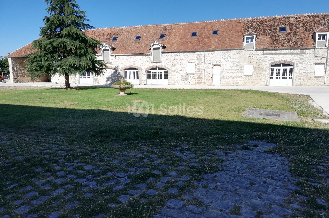 Bâtiment en pierre avec toit en tuiles rouges, grandes fenêtres et pelouse arborée au premier plan.