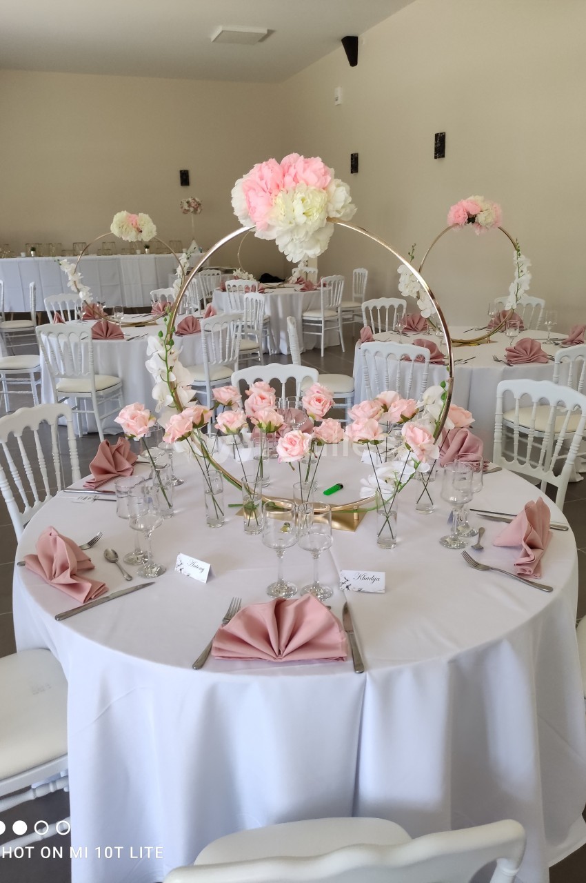 Salle de réception décorée avec des tables rondes, nappes blanches, serviettes roses et centres de table fleuris.