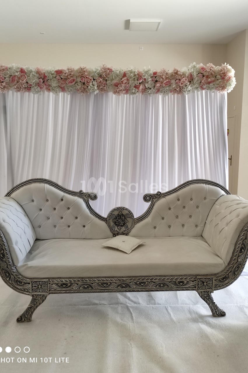 Canapé beige capitonné avec accoudoirs sculptés, placé devant un rideau blanc décoré de fleurs roses et blanches.