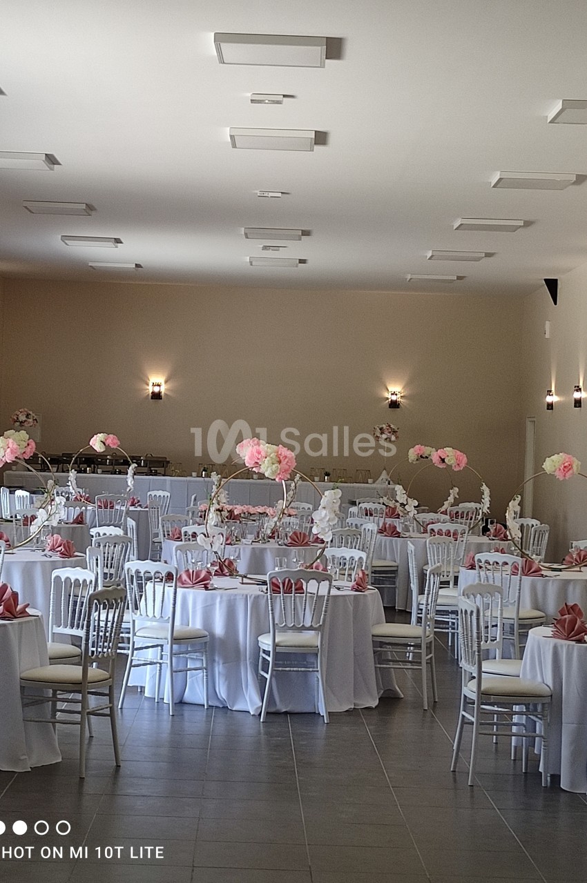 Salle de réception décorée avec des tables rondes, nappes blanches, serviettes roses et centres de table fleuris.