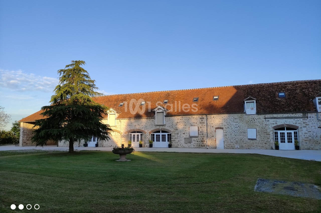 Bâtiment en pierre avec toit en tuiles, grandes fenêtres blanches et pelouse arborée au premier plan.