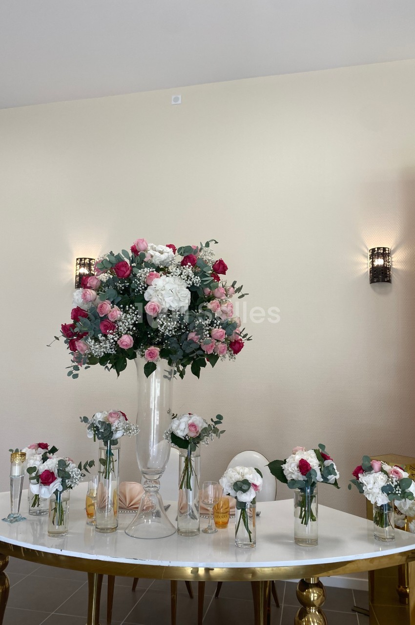 Arrangement floral avec roses roses et blanches dans un vase central, entouré de petits bouquets sur une table blanche.
