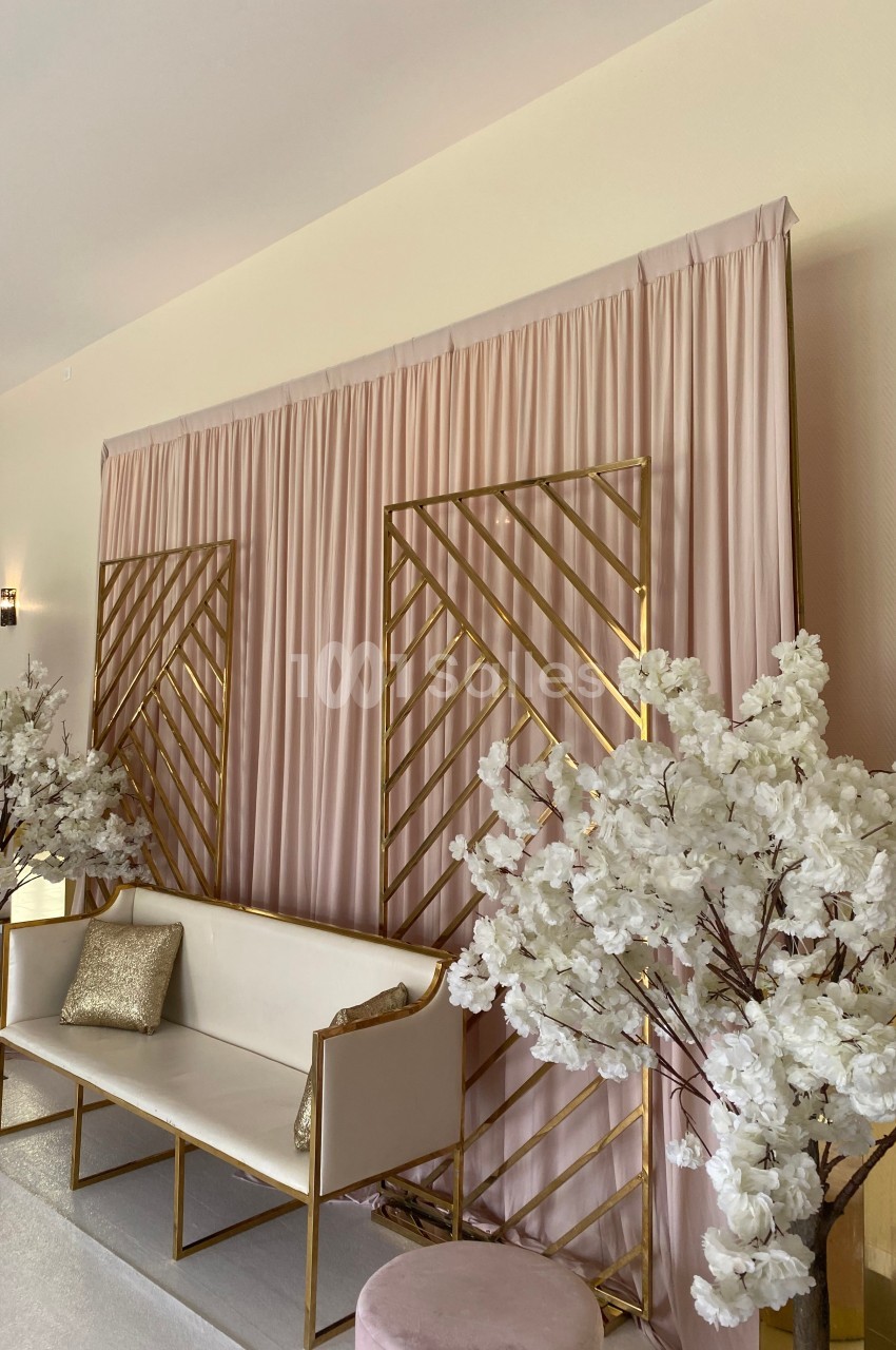 Mur décoratif avec rideaux roses, structures dorées, canapé blanc, coussins dorés et arbres fleuris blancs.